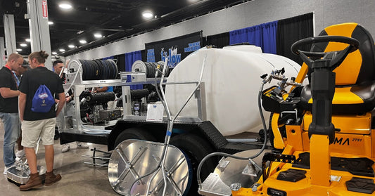 A yellow BEAST riding surface cleaner sits beside a commercial pressure washing trailer and equipment at a trade show.