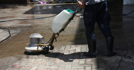 A worker operates a rotary surface cleaning machine on brick pavers, with wet and cleaned sections visible.