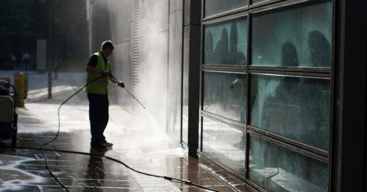 A commercial pressure washing operator cleans a large paved area outside a commercial building with high water flow.